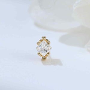 May include: A small gold stud earring featuring a central marquise-cut clear gemstone held by a delicate metal setting with decorative beaded accents along the edges, creating a refined and symmetrical appearance for ear piercing jewelry.