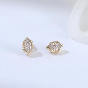 May include: A pair of gold stud earrings, each featuring a marquise-cut clear gemstone set in a pointed metal frame. The earrings are displayed side-by-side, showcasing the elongated diamond-like shape of the stones and the polished gold metal settings.