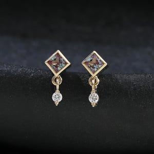 May include: A pair of gold-colored earrings. Each earring features a square-shaped, multi-colored gemstone set within a gold frame. A small, round, clear gemstone dangles below each square. The earrings are displayed on a dark, textured surface.