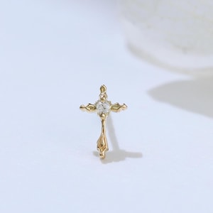 May include: A small gold stud earring shaped like a cross, featuring a single clear gemstone set at the center. The metal has a polished finish with delicate, pointed ends on the horizontal and vertical bars of the cross design.