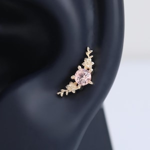 May include: A gold-toned ear stud featuring a central round light pink gemstone flanked by small metallic floral and leaf-shaped accents. The jewelry piece is curved to follow the contour of the ear cartilage, showcasing a delicate botanical design.