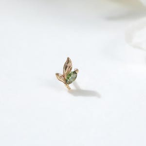 May include: A delicate gold-colored leaf-shaped earring with a small green gemstone. The earring is set against a white background with a blurred white flower in the background. The earring is a small, elegant piece of jewelry.