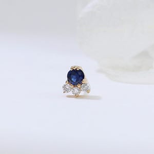 May include: A small, gold-colored stud earring featuring a large, round, dark blue gemstone at the top, with three smaller, clear gemstones below. The earring is set against a soft, white background.
