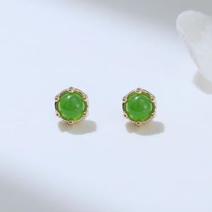 May include: A pair of stud earrings featuring round, vibrant green gemstones set in gold-colored metal. The earrings have a delicate, crown-like design around the gemstones. The background is a soft white, highlighting the jewelry.