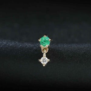 May include: A gold stud earring featuring a round green gemstone set in a prong setting, with a small dangling gold component holding a single clear round gemstone at the base. The earring has a delicate design with a polished gold finish.