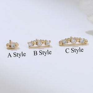 Puede incluir: Tres pendientes de botón dorados con piedras preciosas transparentes. Los pendientes están etiquetados como "A Style", "B Style" y "C Style". Los pendientes se muestran sobre una superficie blanca.