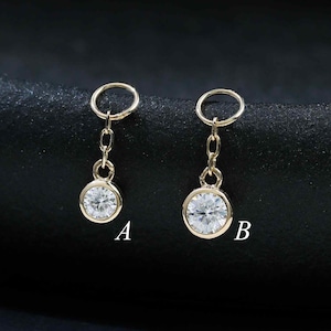 May include: Two gold-toned dangle charms, labeled A and B, each featuring a small circular bezel-set clear gemstone suspended from a delicate chain link and a circular jump ring. The charms are metallic gold in color with clear stones.