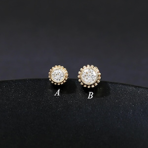 Pode incluir: Dois brincos de botão dourados com uma pedra central transparente e uma borda decorativa com contas. Os brincos estão rotulados A e B. O fundo é de cor escura e lisa.