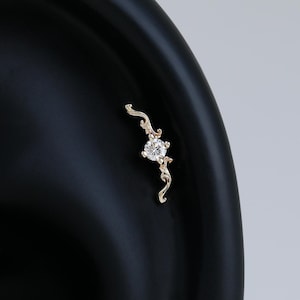 May include: A gold ear piercing stud featuring a central round clear gemstone set within a curved, vine-like metal frame. The delicate gold structure extends outward from the center stone in two directions, creating a slender, elongated shape.