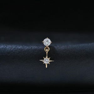 May include: A gold stud earring featuring a small round clear gemstone at the top, connected by a thin gold link to a dangling star-shaped charm with a central clear gemstone. The design incorporates delicate metal prongs and fine detailing.