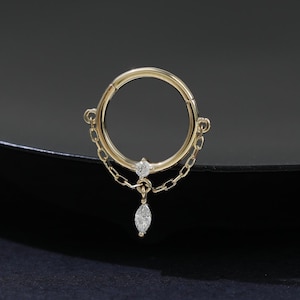 May include: A gold-colored hinged nose ring with a dangling chain and a teardrop-shaped gem. The ring has a circular design with a small gem at the top and a chain that connects to a teardrop-shaped gem. The jewelry is set against a dark background.