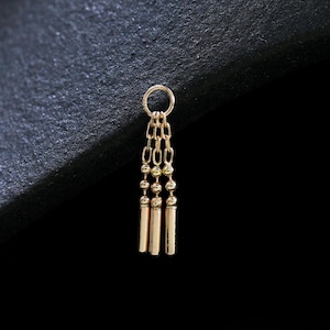 Peut inclure: Un petit charme de boucle d'oreille de couleur or avec un design pendant. Le charme présente un dessus circulaire relié à trois barres cylindriques verticales. Les barres sont reliées par de petites chaînes et des perles. La boucle d'oreille est posée sur un fond sombre.