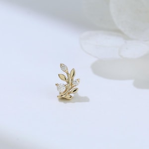 May include: A gold-coloured stud earring shaped like a delicate leaf, adorned with small, sparkling clear gemstones. The earring features a nature-inspired design, ideal for adding a touch of elegance to any outfit.