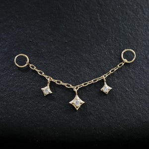 May include: A delicate gold-coloured body chain with three diamond-shaped pendants. The chain features two small rings on each end and a central pendant. The pendants are adorned with small, sparkling gemstones, creating a subtle, elegant design.