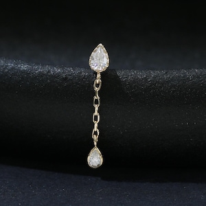 May include: A gold earring featuring a small pear-shaped clear gemstone at the top, connected by a delicate gold chain to a second, smaller pear-shaped clear gemstone dangling at the bottom, creating a vertical drop design with two stones.