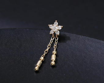 May include: A gold-colored earring featuring a small, five-petal flower design with clear, sparkling stones. Two delicate chains dangle from the flower, each ending with a small, cylindrical bead. The earring is set against a dark background.