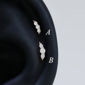Pode incluir: Dois piercings dourados para a orelha, cada um com três pedras preciosas redondas e transparentes. Os piercings estão colocados num modelo de orelha preto, um acima do outro. As pedras preciosas estão dispostas numa linha curva, criando um visual delicado e elegante.