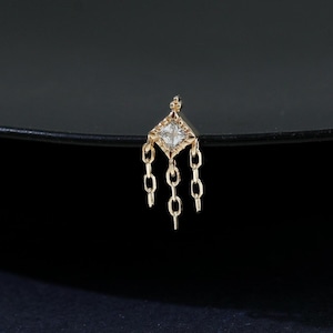 Puede incluir: Un pequeño pendiente dorado con una parte superior en forma de diamante y tres cadenas colgantes. El pendiente se encuentra sobre una superficie oscura y reflectante, resaltando su delicado diseño y sus intrincados detalles.