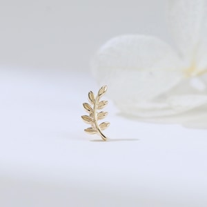 May include: A small, gold-colored earring in the shape of a delicate leaf. The earring is positioned on a white surface, with a blurred white flower in the background. The earring's design features intricate details, suggesting a nature-inspired theme.