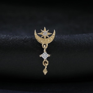 May include: A gold-coloured celestial-themed earring. The earring features a crescent moon with a star, a dangling square-shaped gem, and a teardrop-shaped charm. The earring is designed with small, sparkling accents.