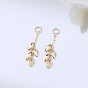 May include: A pair of gold-colored leaf-shaped earrings. Each earring features a chain with three leaf charms and a small loop at the top. The earrings are displayed on a white background with soft lighting.
