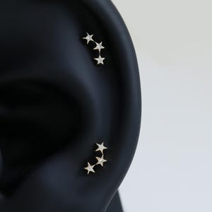 以下が含まれることがあります： 黒い耳のモデルは、小さな金色の星型のイヤリングを2組表示しています。3つの星の1組はヘリックスに沿って配置され、3つの星の別の組は耳たぶにあり、天体のテーマを示しています。