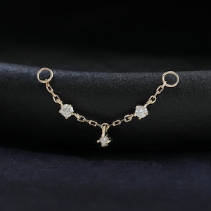Könnte beinhalten: Eine zarte Goldkette mit drei klaren, funkelnden Edelsteinen und einem kleinen sternförmigen Anhänger. Die Kette hat an beiden Enden zwei kleine Schlaufen, die zum Piercen bestimmt sind. Der Schmuck ist vor einem dunklen Hintergrund platziert.