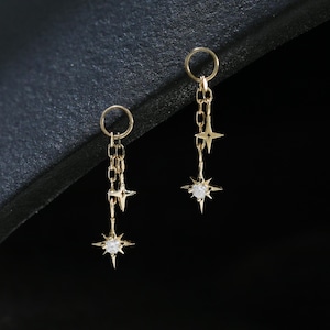 Puede incluir: Un par de pendientes colgantes dorados. Cada pendiente presenta un pequeño aro, una cadena y tres colgantes en forma de estrella. El colgante de estrella inferior tiene una pequeña gema transparente en el centro. Los pendientes se muestran sobre un fondo oscuro.