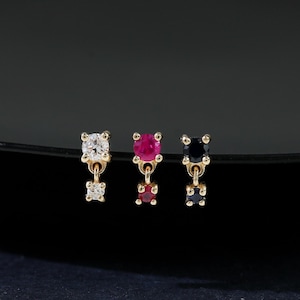 May include: Three gold earrings with different colored gemstones. One earring has a clear gemstone, the second has a red gemstone, and the third has a black gemstone. Each earring has a small, dangling gemstone below the main stone.