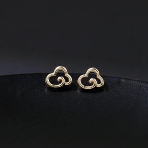 Könnte beinhalten: Ein Paar kleine, goldfarbene Ohrstecker in Wolkenform. Die Ohrringe liegen auf einer schwarzen Oberfläche mit dunklem Hintergrund. Die Ohrringe haben ein schlichtes, minimalistisches Design.