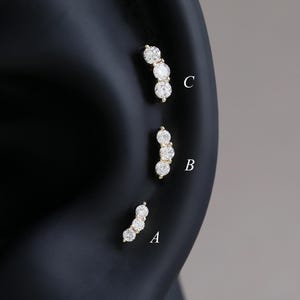 Può includere: Tre orecchini curvi color oro, ciascuno con tre gemme rotonde e trasparenti. Gli orecchini sono esposti su un modello di orecchio nero, con ogni orecchino etichettato A, B e C. Le gemme sono incastonate vicine.