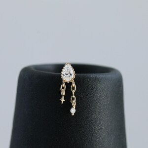 May include: A gold-colored earring with a pear-shaped, clear gemstone at the top. The earring has a chain design with a small star and a round, clear gemstone dangling from the bottom. The earring is displayed on a black, cylindrical object.
