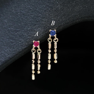 May include: A pair of gold-colored dangle earrings. One earring features a red square gemstone, while the other has a blue square gemstone. Each earring has three dangling chains with small, rectangular gold beads.