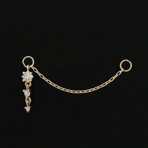 May include: A delicate gold-colored ear chain with a dangling floral design and three small, sparkling gems. The chain connects to a small hoop on the other end. The background is solid black.