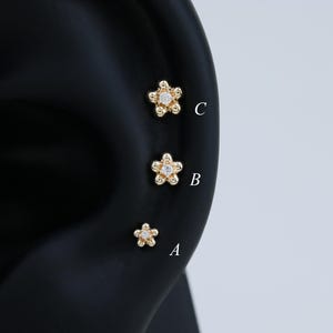 May include: Three gold-colored flower-shaped earrings with small, clear gemstones in the center are displayed on a black ear model. The earrings are labeled A, B, and C, with C being the largest and A the smallest.