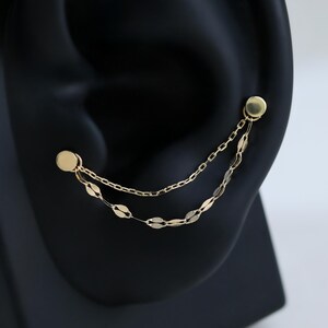 May include: A gold double chain earring featuring two small circular studs connected by a delicate cable chain and a secondary chain with flat, elongated oval links. The earring is designed to span across the earlobe with a polished finish.