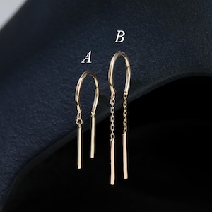 May include: Two pairs of gold-colored threader earrings. One pair features a simple bar design, while the other has a chain detail. The earrings are displayed against a dark background, highlighting their delicate design.