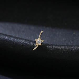 May include: A delicate gold nose ring with a star-shaped charm, set with a single clear gemstone. The curved post extends from the star, creating a graceful design. The jewellery is displayed against a dark, textured background.