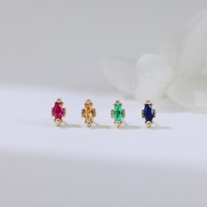 May include: Four gold stud earrings, each featuring a different colored gemstone: red, yellow, green, and blue. The gemstones are marquise-cut and set in a gold bezel. The earrings are displayed on a white surface.
