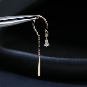 May include: A single gold earring with a chain and a small, pear-shaped gemstone. The earring has a straight bar at the bottom and is held by tweezers against a dark background. The jewellery is elegant and minimalist in design.