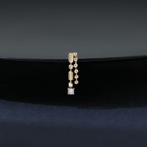 May include: A single gold earring with a dangling design. The earring features a small, square-cut diamond at the bottom, with rectangular and spherical gold elements suspended above. The earring is set against a dark background.