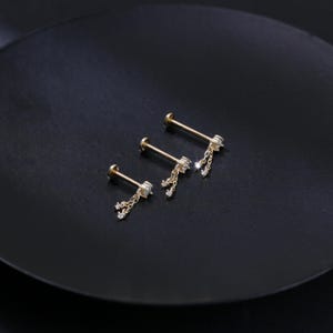 May include: Three gold-colored lip or ear studs with dangling chains and small, clear gemstones. The studs are arranged on a dark, matte surface, showcasing their delicate design and sparkle. The jewelry is designed for body piercing.