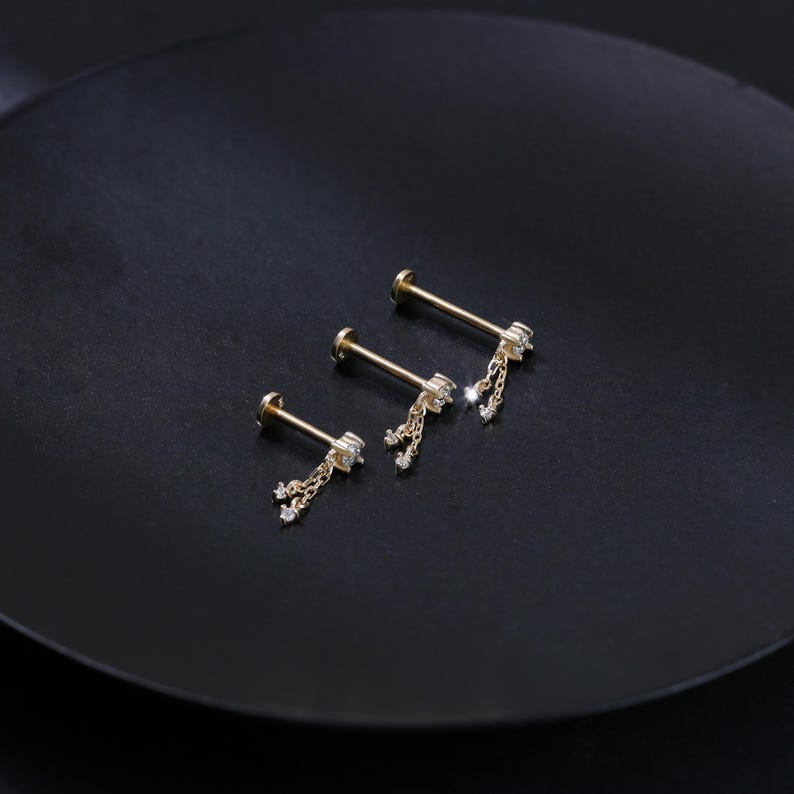 May include: Three gold-colored lip or ear studs with dangling chains and small, clear gemstones. The studs are arranged on a dark, matte surface, showcasing their delicate design and sparkle. The jewelry is designed for body piercing.