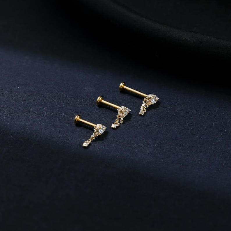 Puede incluir: Tres labrets dorados con un dise&ntilde;o colgante y ornamentado. Cada labret presenta una peque&ntilde;a gema transparente en el centro del dise&ntilde;o y un poste para la inserci&oacute;n. Los labrets est&aacute;n dispuestos en diagonal sobre un fondo oscuro.