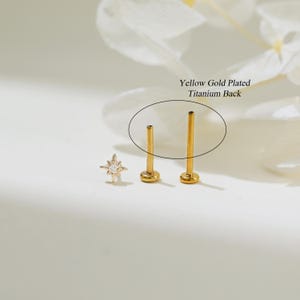 May include: Close-up of a gold star-shaped stud earring with a single clear gemstone in the center. The earring is shown next to two gold-plated titanium earring backs. The text "Yellow Gold Plated Titanium Back" is visible in the image.