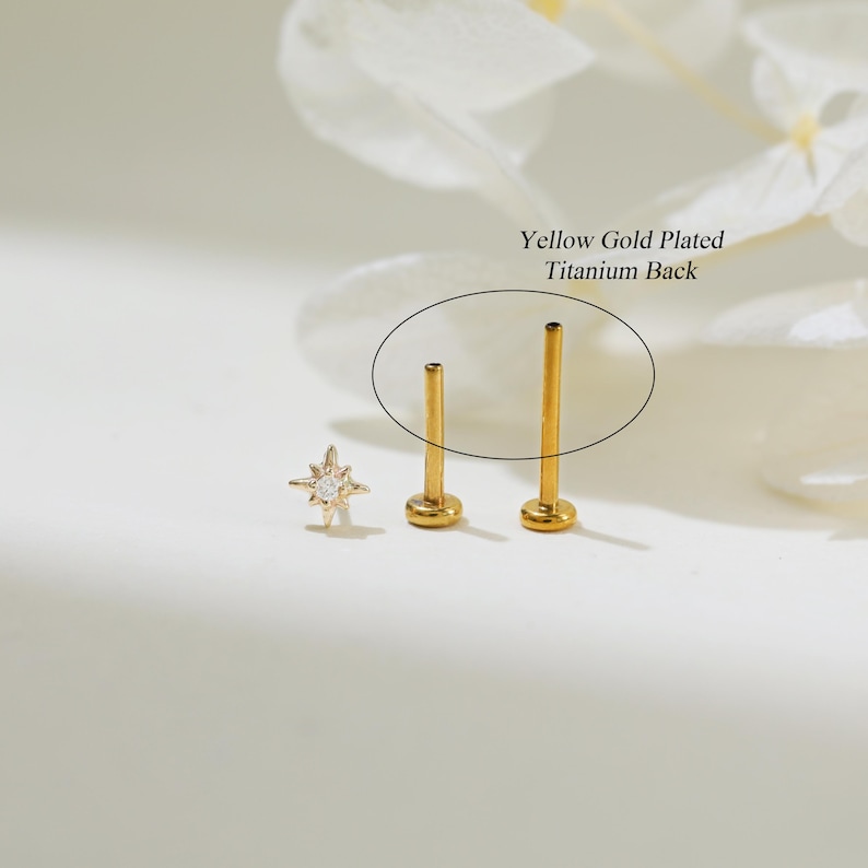 May include: Close-up of a gold star-shaped stud earring with a single clear gemstone in the center. The earring is shown next to two gold-plated titanium earring backs. The text "Yellow Gold Plated Titanium Back" is visible in the image.