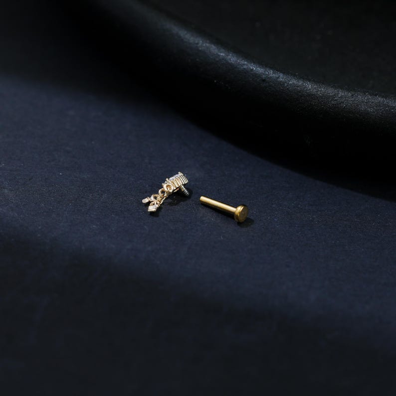 Puede incluir: Un labret de color dorado y un poste de pendiente separado. El labret presenta un peque&ntilde;o dije decorativo con m&uacute;ltiples acentos claros y brillantes. El poste del pendiente tiene un extremo plano y redondo.