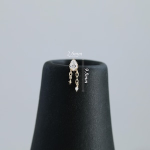 Puede incluir: Un pendiente dorado con una gema transparente en forma de pera y detalles de cadena colgante. El pendiente mide 9,8 mm de largo y 2,6 mm de ancho. Presenta una estrella y una peque&ntilde;a gema redonda al final de las cadenas.