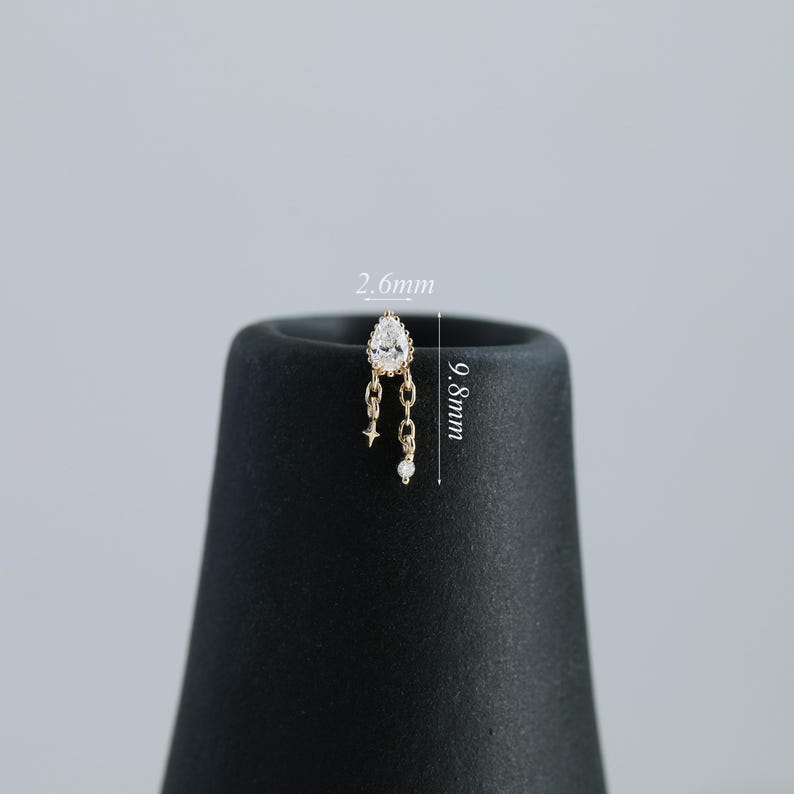 Puede incluir: Un pendiente dorado con una gema transparente en forma de pera y detalles de cadena colgante. El pendiente mide 9,8 mm de largo y 2,6 mm de ancho. Presenta una estrella y una peque&ntilde;a gema redonda al final de las cadenas.
