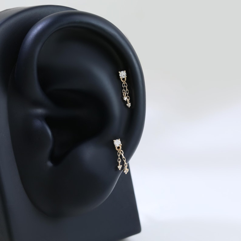 May include: Two gold-colored earrings with a dangling design are displayed on a black ear model. Each earring features a square top with a central clear stone and a chain of smaller stones. The earrings are designed for ear piercings.